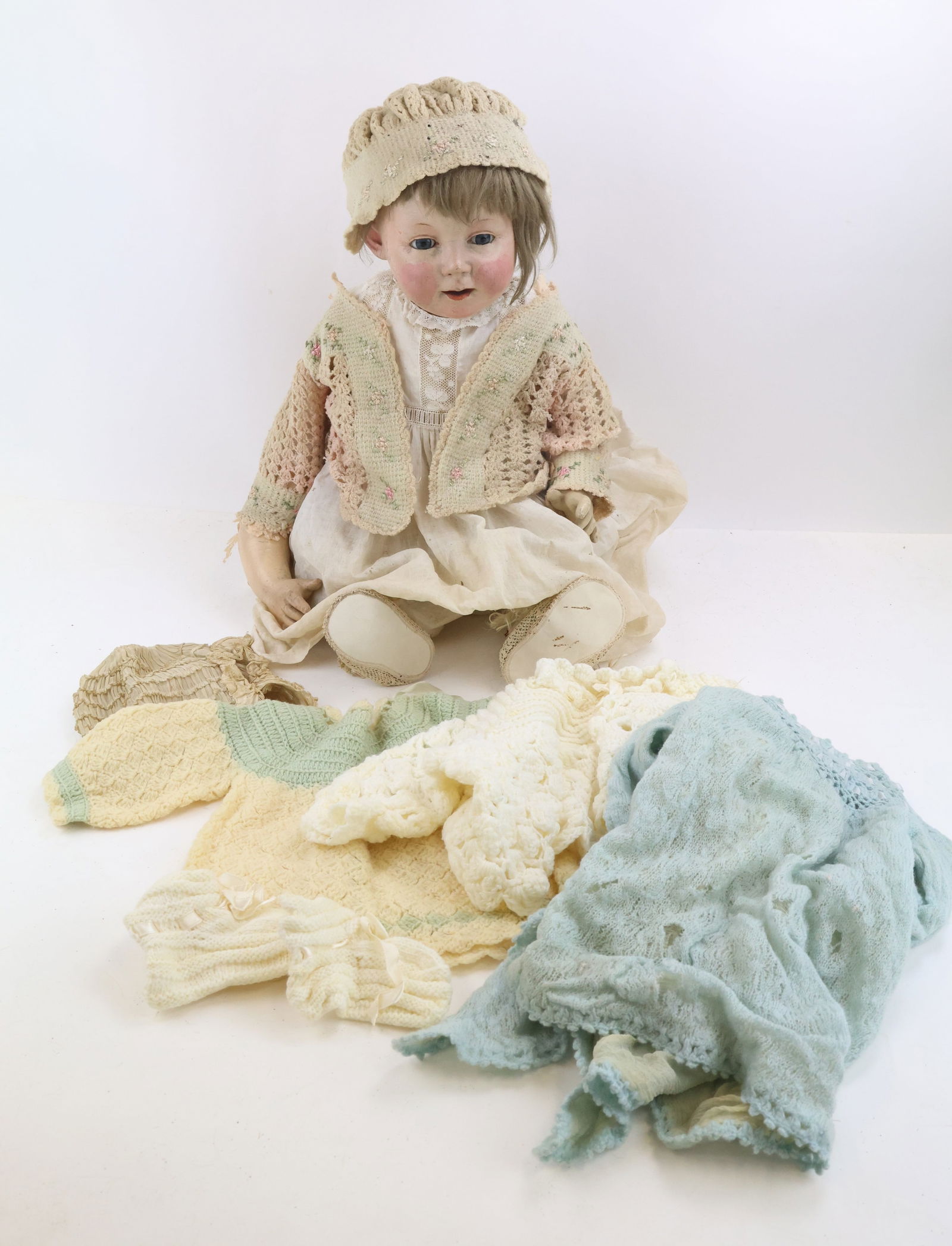 Sitting Papier Mache Baby Doll: With nodding head and stationary glass eyes, mohair wig, unmarked, wearing antique clothing and shoes with additional garments. Age loss to wig, clothing, and body; repaint on face; arm detached. 18"