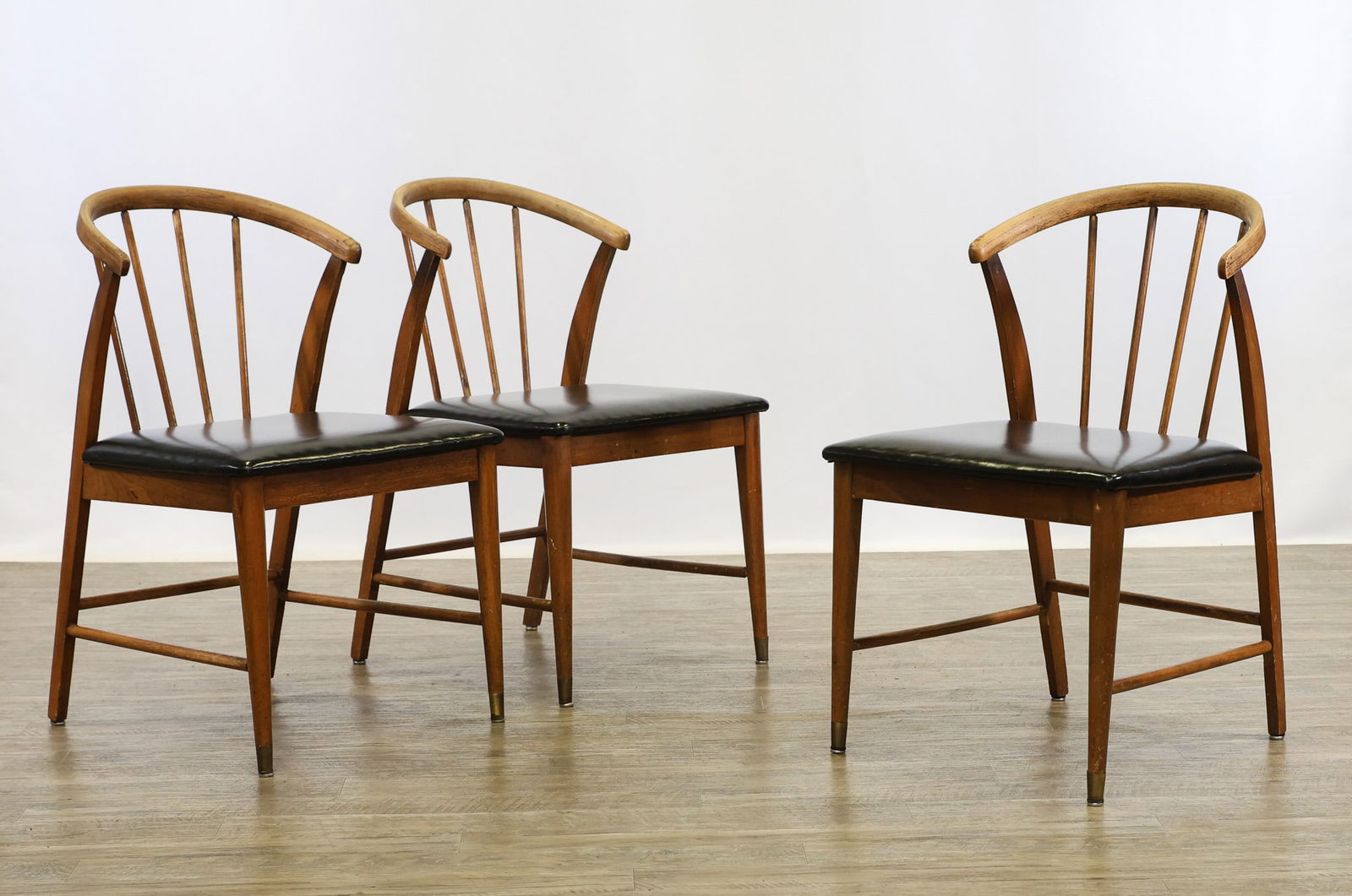 Spindle Back Dining Chairs: Walnut dining chairs with curved spindle backs, tapered and slightly bent square stiles, tapered round front legs with brass caps, round back and side stretchers, and black vinyl seats. Finish loss. (