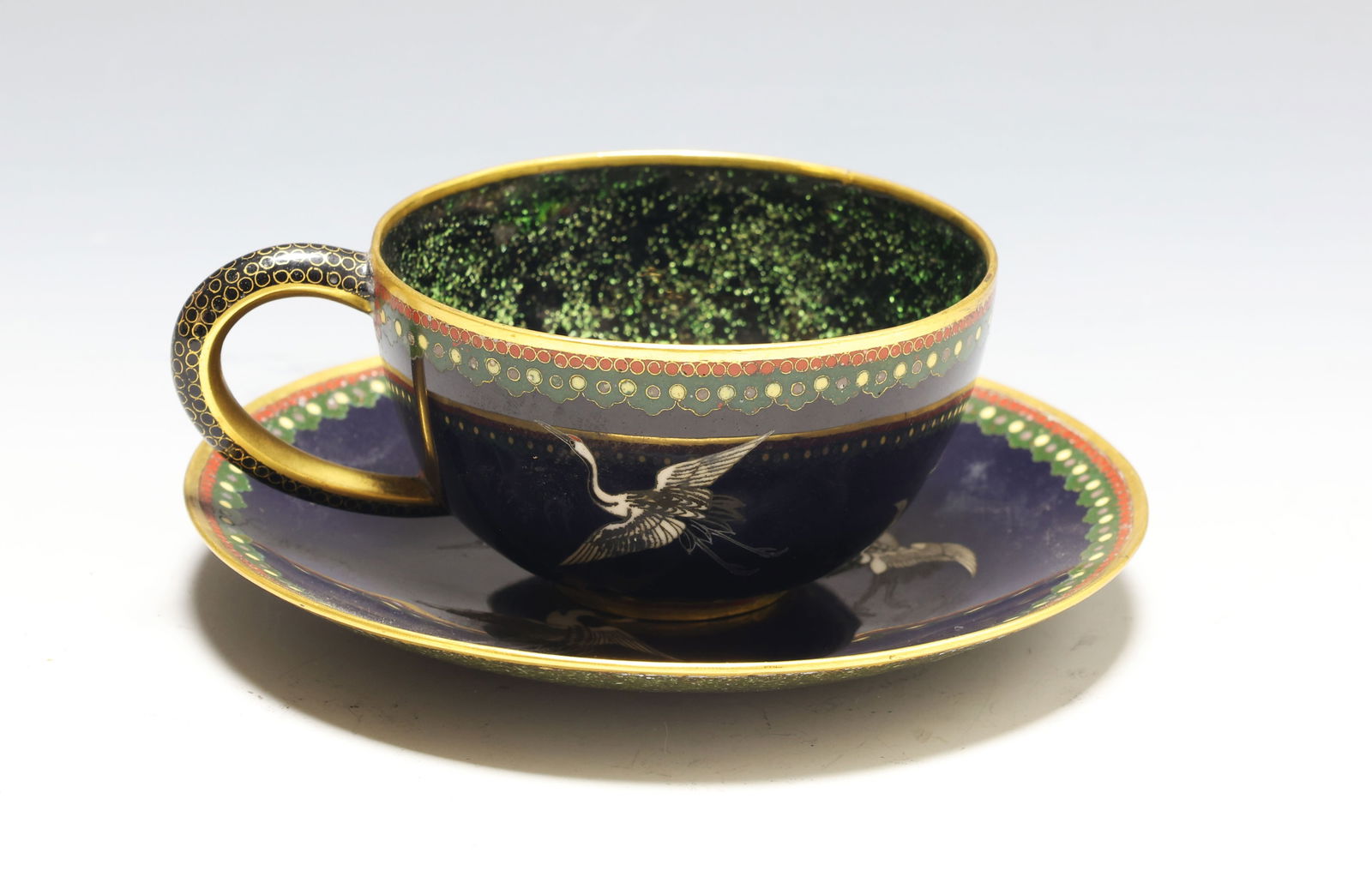 Japanese Goldstone Cloisonne Cup and Saucer: Meiji - Showa era parcel gilt silver wire cloisonne teacup and saucer, depicting cranes on a black ground, with stylized rosette and fish roe borders, and wire scroll and goldstone accents. Unmarked.