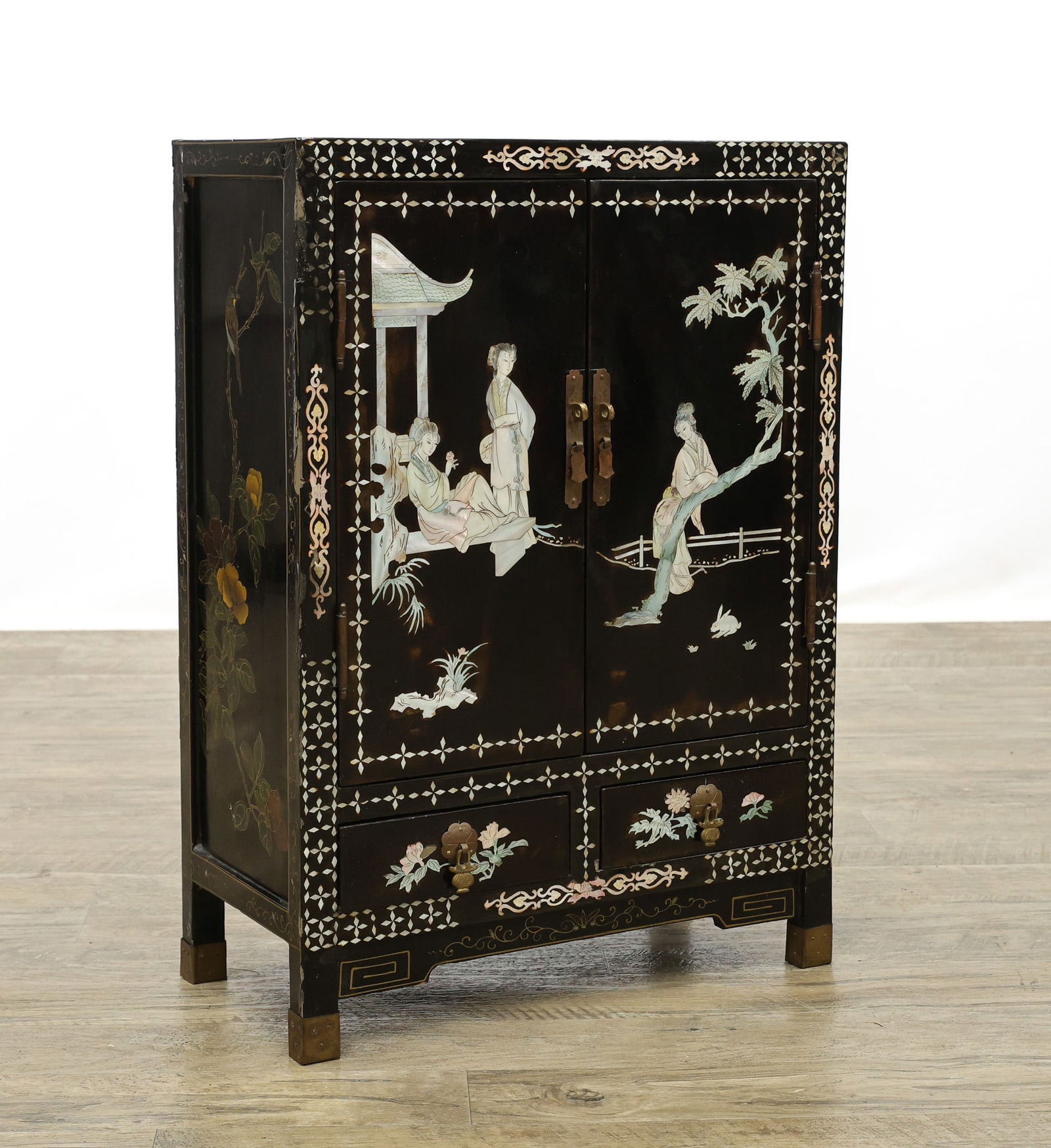 Chinese Export Mother of Pearl Inlaid Lacquer Cabinet: Black lacquer case inlaid with mother-of-pearl court figures in garden scenes, geometric borders and floral motifs, paneled doors over two drawers, brass hardware, raised on bracket feet. 26"h x 18.5"
