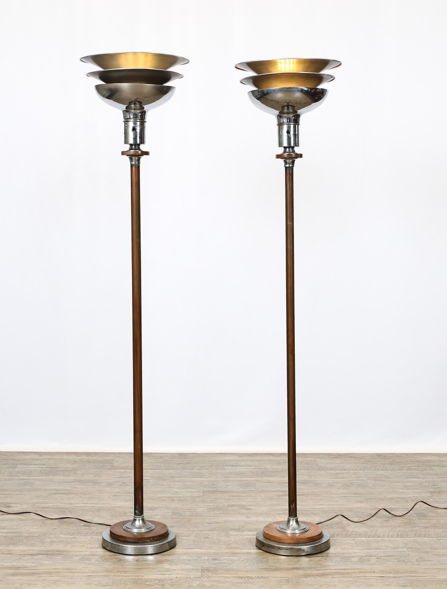 Gilbert Rohde Torchieres: Art Deco pair of torchieres, with copper stems, chromed aluminum bases and tiered tops with walnut accents. Unmarked. Newer wiring; both in working condition. Bends to the metal; pitting to the base;