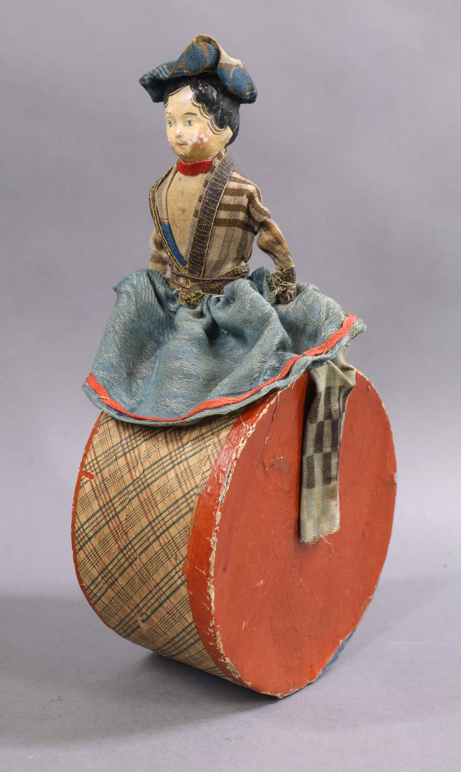 TOYS - ANTIQUE.: Early German papier mache "Roly" Toy. Doll sitting on a drum like base. Greiner type papier mache head with painted features. Wire armature arms to her side, legs straddling over the drum. Clothing
