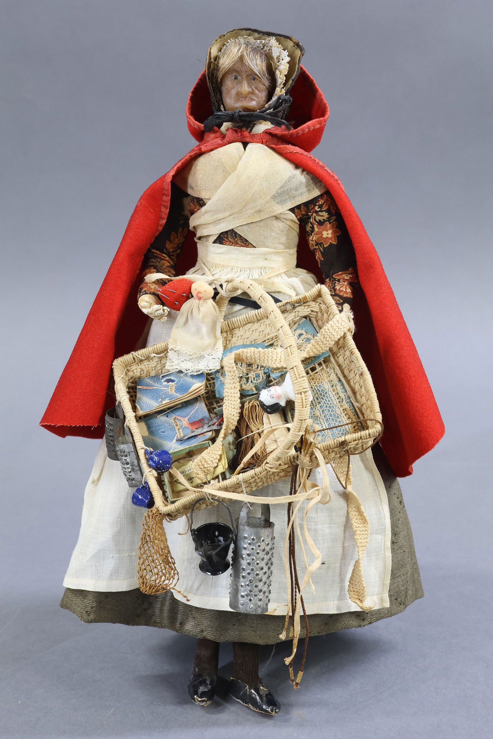 ANTIQUE PEDDLER.: 11" Wax Head Lady Peddler Doll. England, Mid 19th c. Painted features. Costume of red suede hooded cape, black silk bonnet. She is carrying a basket of sewing and metal cooking wares. Mini China