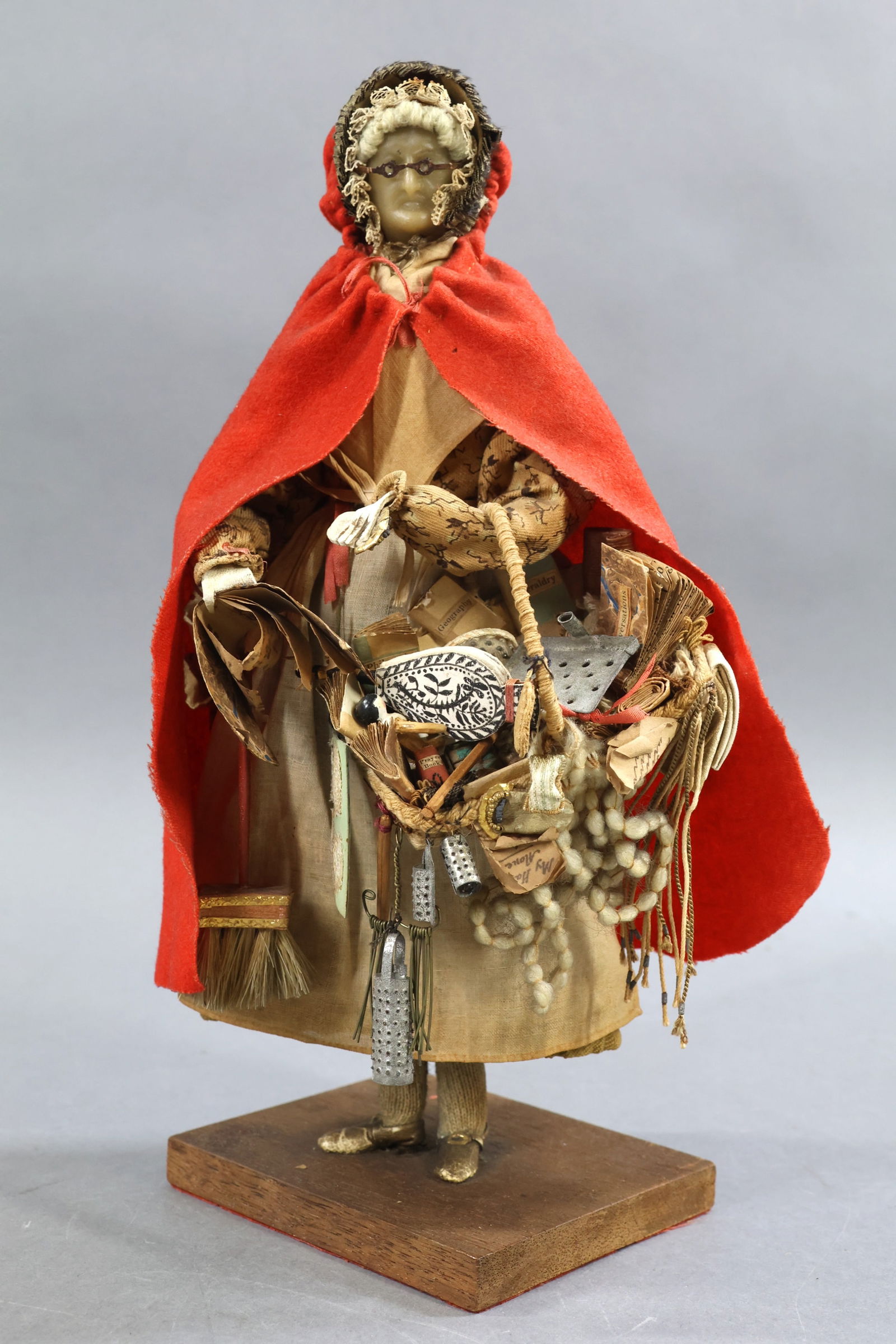 ANTIQUE PEDDLER.: 11" Wax Head Lady Peddler Doll. England, Mid 19th c. Painted features. Spectacles. Costume of red cloth hooded cape, brown silk bonnet with ruching on front. She is carrying a basket of misc. wares