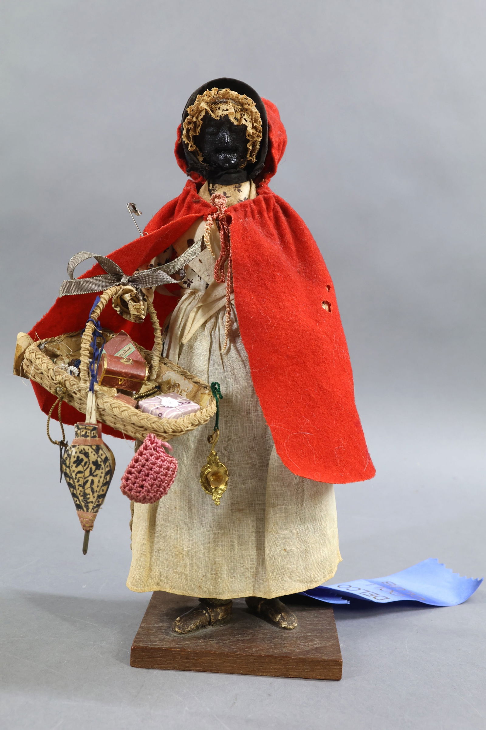 ANTIQUE PEDDLER.: 11" Wax? head Lady Peddler Doll. Mid 19th c, England. Costume of red wool hooded cape, black bonnet with rows of lace underneath. She is carrying a basket of misc. wares. She is mounted on a wooden