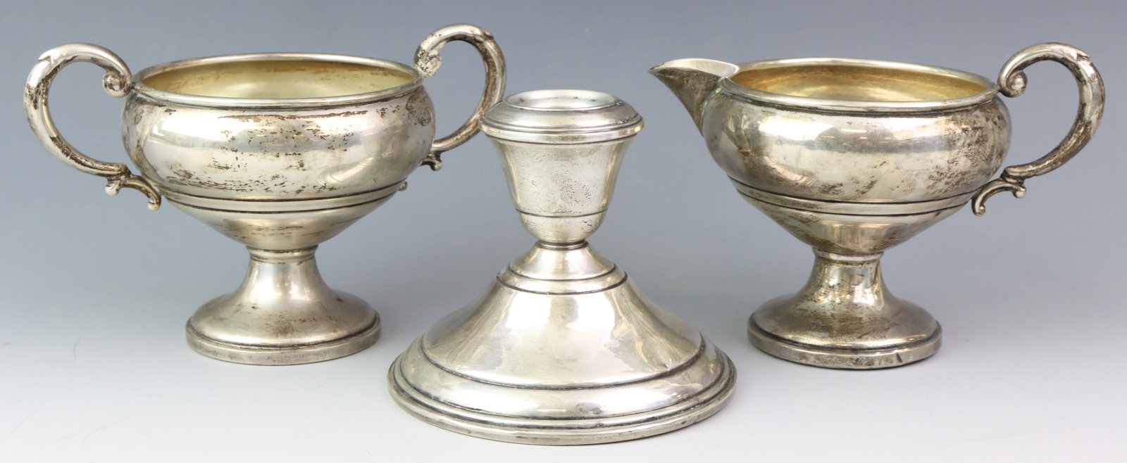 Mueck-Carey and Schweitzer Sterling Silver Tablewares: American sterling silver weighted pieces with complementing patterns, including a Mueck-Carey creamer and sugar bowl set, and a Schweitzer candlestick. (3) pieces. Sugar 3.5”h x 5.5”w x 3.