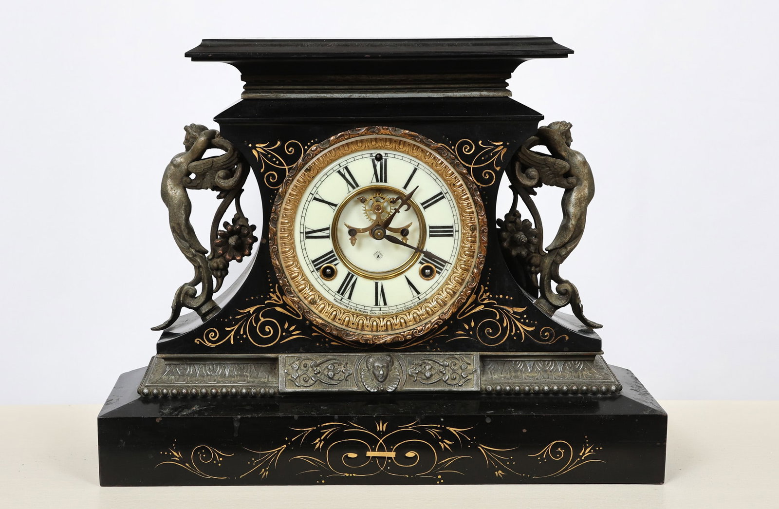 Ansonia Neo-Classical Style Mantel Clock: Black marble case having concave sides with cast metal cherub handles, gadrooned symbolic and figural molding, and engraved foliate motifs. Enameled dial with brass 8-day spring driven works. 12"h x