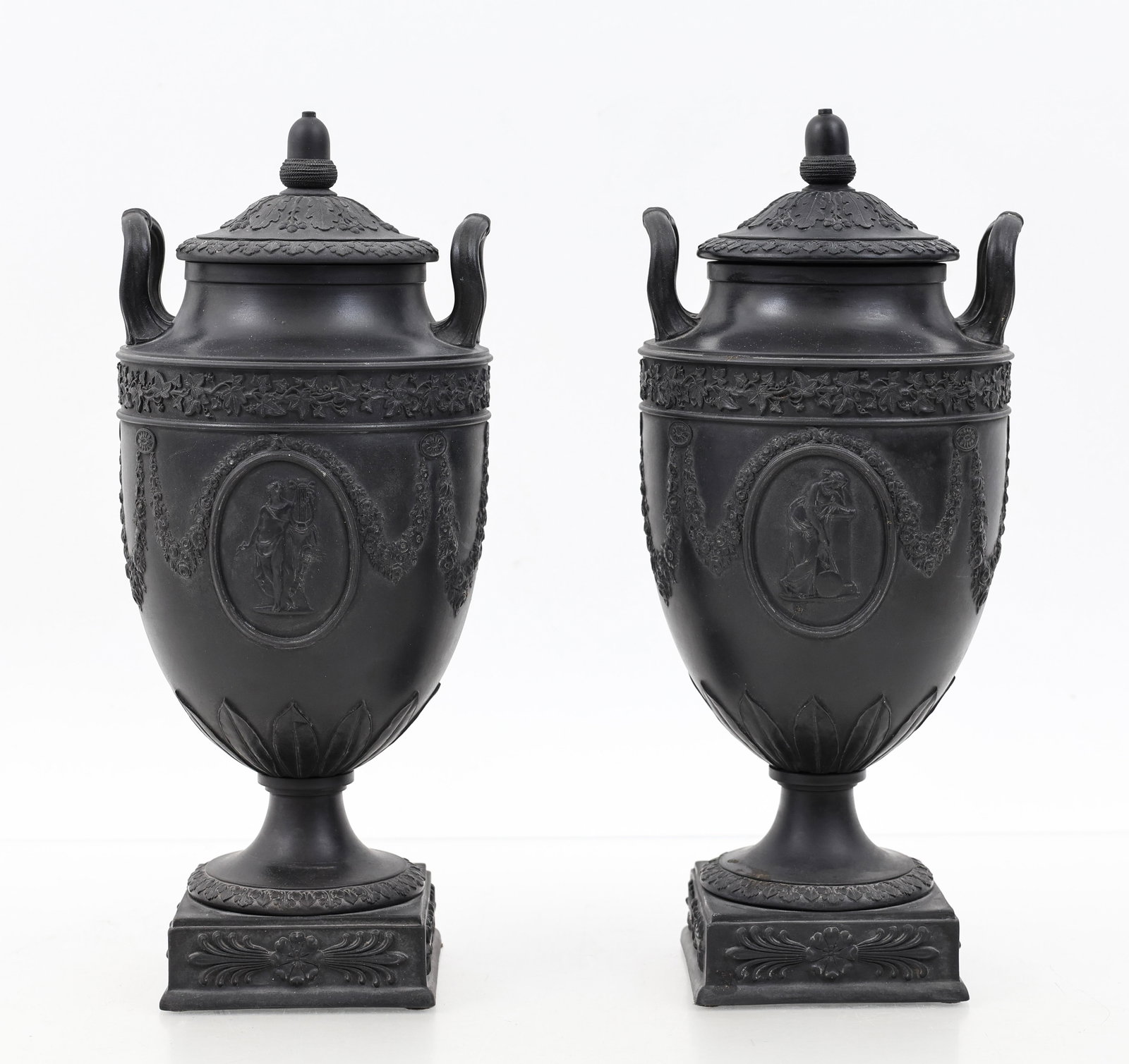 Wedgwood Black Basalt Urns: British stoneware pair of Neoclassical urns, decorated with relief figures in medallions accented by festoons, featuring oak leaf and rosette motifs, handles, and covers with acorn finials. Circa