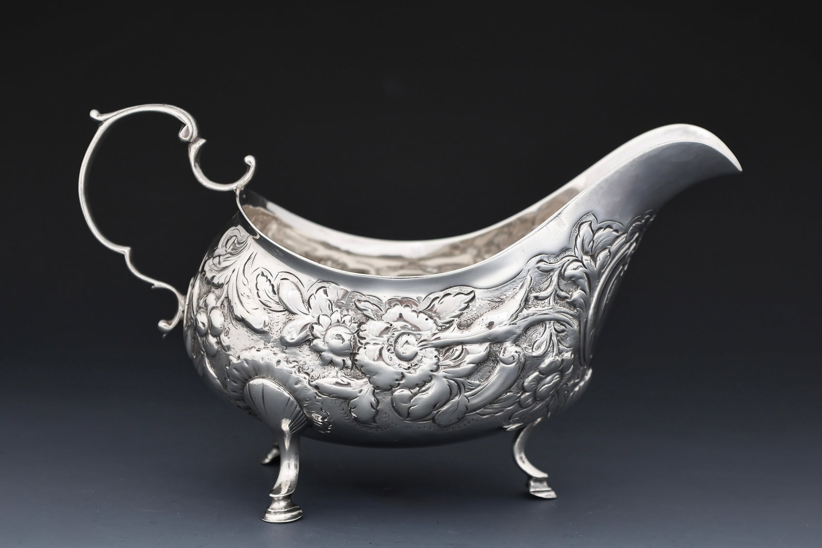 George West Georgian Irish Sterling Silver Sauceboat: George III sterling silver sauceboat with repousse flower, grape, wings, and acanthus decoration, a scroll handle, and raised on shell and hoof feet. Dublin, 1795, by George West. 4.25"h x 7.25"w. 6.5