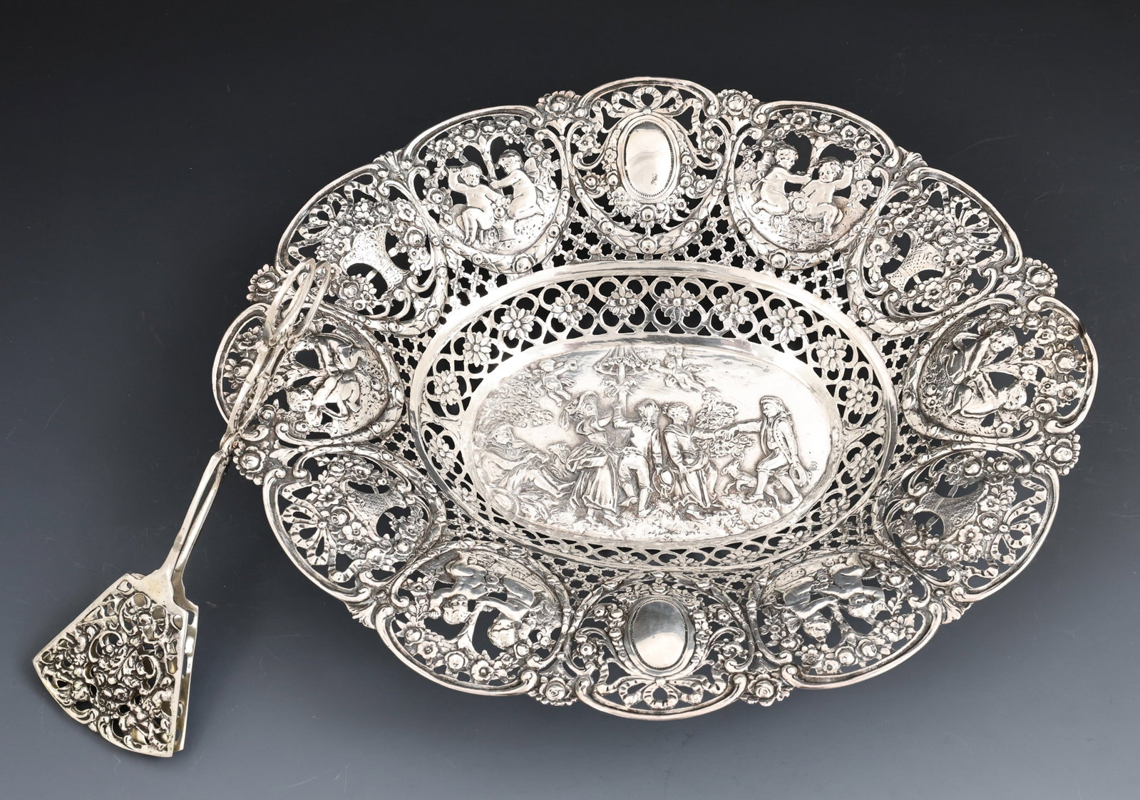 German Silver Bonbon Basket and Tongs: German 800 silver bonbon basket with reticulated openwork and repousse decoration depicting couples and animals dancing in a garden setting, putti, flower bouquets, and rosettes, with an unidentified