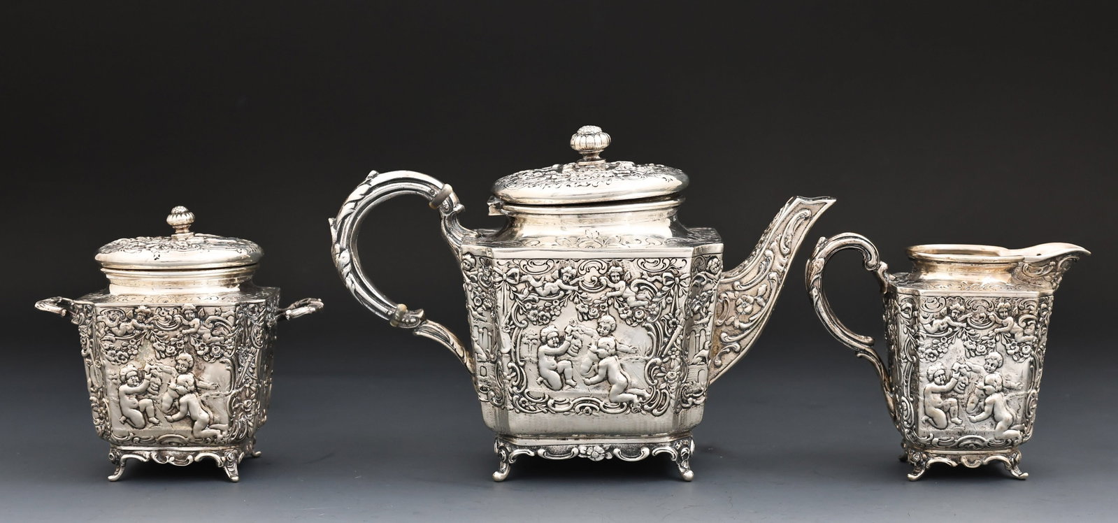Storck and Sinsheimer Silver Tea Service: German Hanau 800 silver service, including a teapot, a creamer, and a covered sugar bowl, with repousse decoration depicting putti in a garden of flowers with animals and Classical buildings. Marked