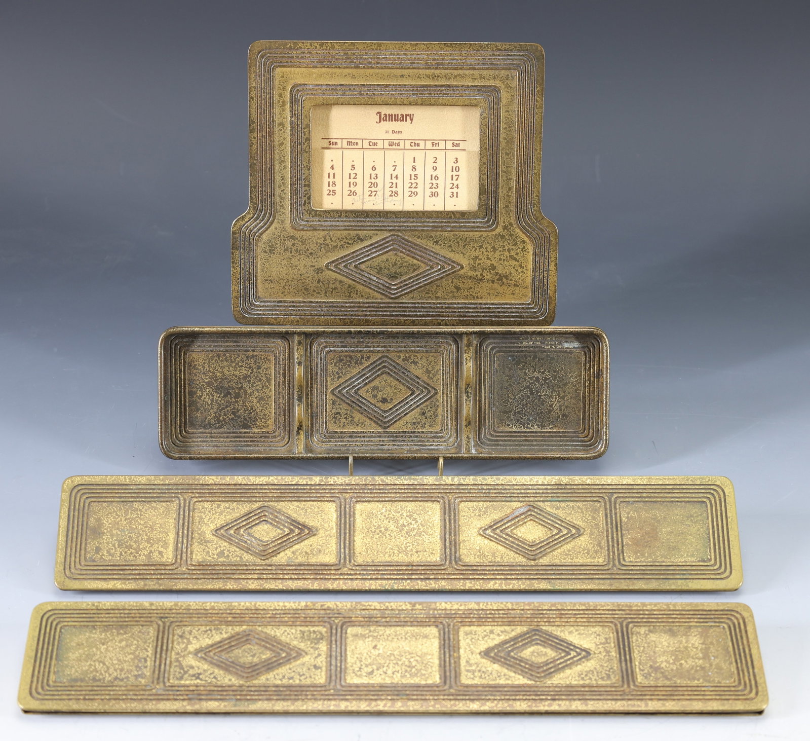 Tiffany Studios "Graduate" Desk Set Accessories: Arts and Crafts gilt bronze accessories, including calendar no. 1804 (date cards incomplete), pen tray no. 1798, and (2) blotter holders 1795. (4) pieces. Calendar 5.5"h x 6.25"w; tray 8.5"w x 2.5"d;