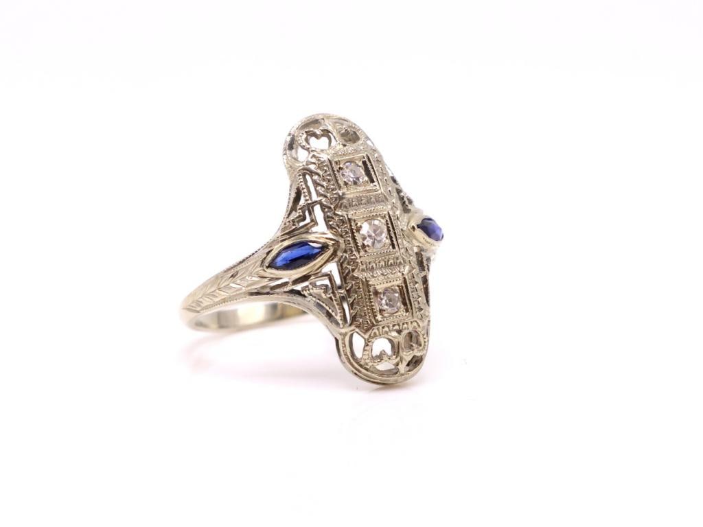 18k WG Diamond Sapphire Ring: 18K white gold ring with three round cut diamonds totaling approximately .10 ctw within an openwork filigree mount, accented by two marquise cut sapphires. Size 5.75, 2.1 dwt.