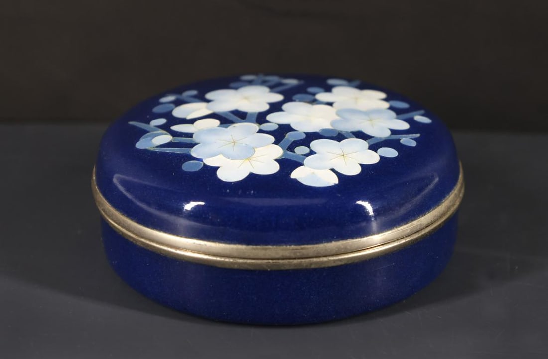 Ando Japanese Cloisonne Box: Showa era silver wire cloisonne round box with silver plated mounts, decorated with plum blossoms on a lapis blue ground, and featuring a tufted red velvet interior. Marked to the foot for Ando. Some 
