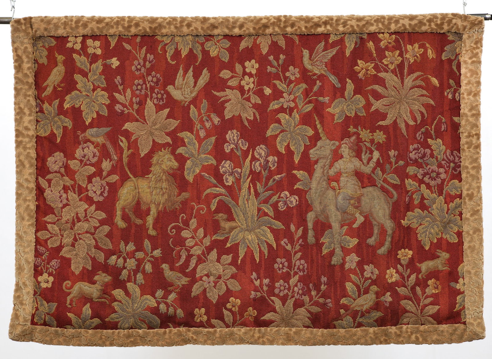 French Unicorn and Lion Tapestry: Handwoven forest pictorial of a unicorn, figural, lion, and aviary motifs in a red ground with a light brown velvet foliate border, and an iron fluer de lis hanging rod. 44"h x 61"w, hanging piece