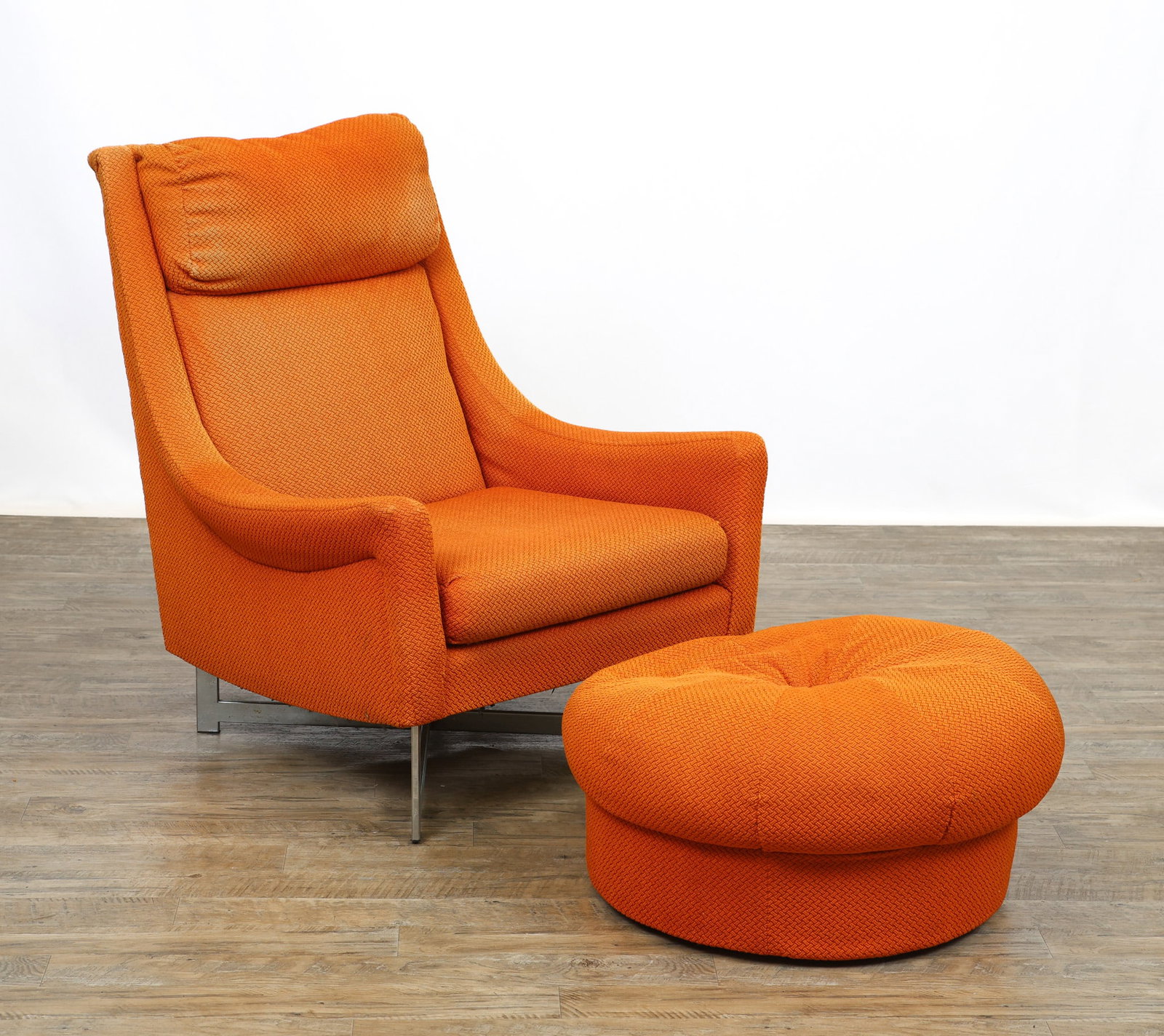 Lounge Chair and Ottoman: Lounge chair with outswept arms, textured burnt orange upholstery, and raised on a chrome steel bar X-form base, with a tufted round pouff ottoman. Fading to the upholstery, some pitting to the