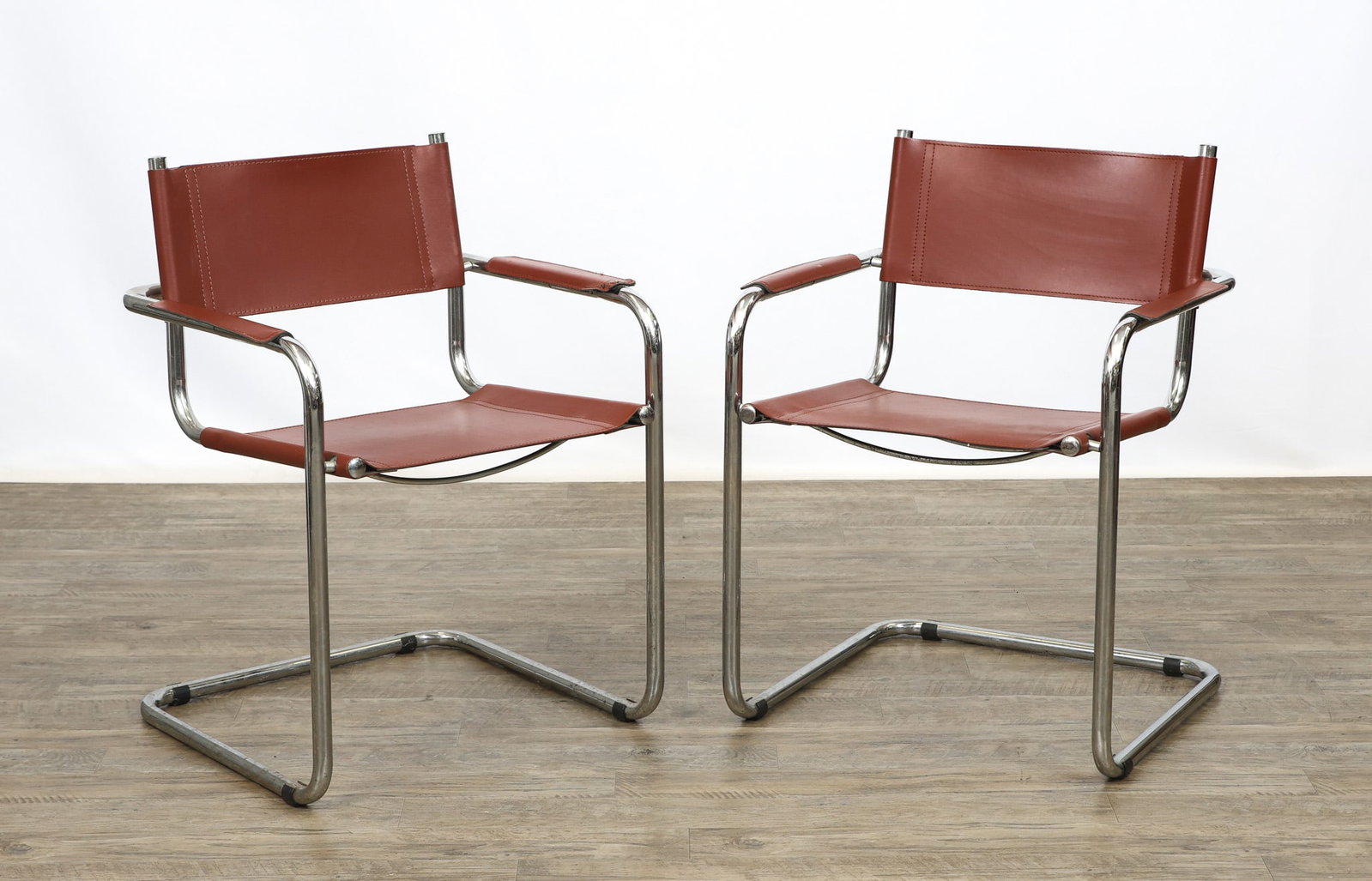 Cantilever Chairs: Tubular chrome pair of cantilever chairs with rust red leather seats, backs, and arms. Some wear to the leather including scuffs/scratches, and loose thread on an arm. (2) chairs. 31"h x 23.5"w x 22"d