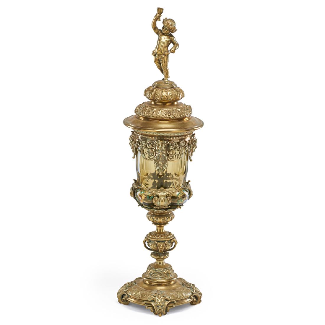GILT BRONZE AND ENAMELED GLASS POKAL: Large amber glass pokal with foliate enamel decoration resting in gilt bronze mounts, adorned with various male heads and a finial of a cherub drinking wine, Continental, late 19th c. 25" x 6 1/2" dia