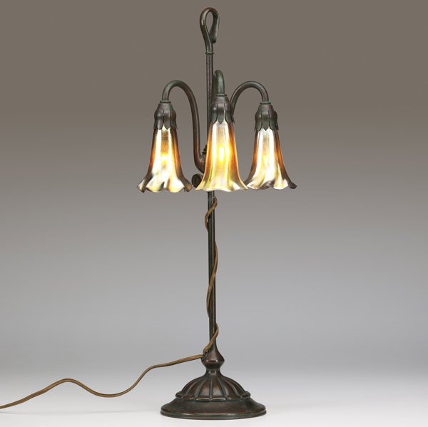 TIFFANY STUDIOS Three-Lily lamp: TIFFANY STUDIOS Three-light Lily table lamp, with gold Favrile shades on an adjustable floriform bronze base with verdigris patina. From a local estate, we believe this piece to be in mint original co