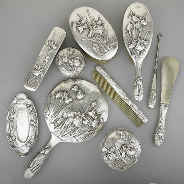 JAPANESE EXPORT SILVER VANITY SET, ca. 1900: Ten pieces with embossed iris motifs on stippled ground: two round covered boxes, 4" x 3"; pin tray, 6"; button hooks; shoe horn, 8 3/4"; three brushes, longest 9 1/4"; comb, 7 1/4"; hand mirror, 11".