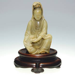 Oriental soapstone carving of Guan Yin, 4 1/2",sta: Oriental soapstone carving of a seated Quan Yin caressing a Foo puppy. Height 4 3/8 inches. Her hair highlighted in black enamel, her robe engraved with designs. Height 4 1/2 inches. A carved wood sta