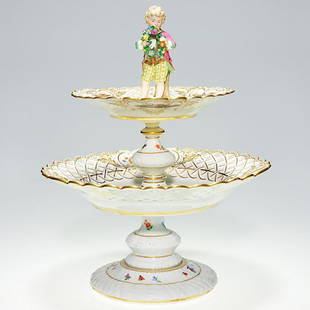 Meissen porcelain 2 tier compote, boy, 15": Meissen two tier porcelain compote with a figure of a young barefoot lad grasping a floral wreath wearing knickers and pink coat. The tiers he guards are pierced and decorated with colorful flowers an