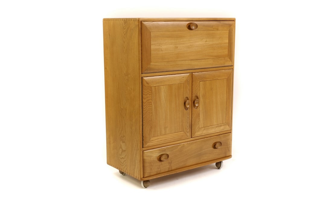 An Ercol beech cocktail cabinet: An Ercol beech cocktail cabinet An Ercol beech cocktail cabinet, c.1960s, the fall front enclosing a split interior with two adjustable shelves, over a two-door cupboard with a single shelf and lower