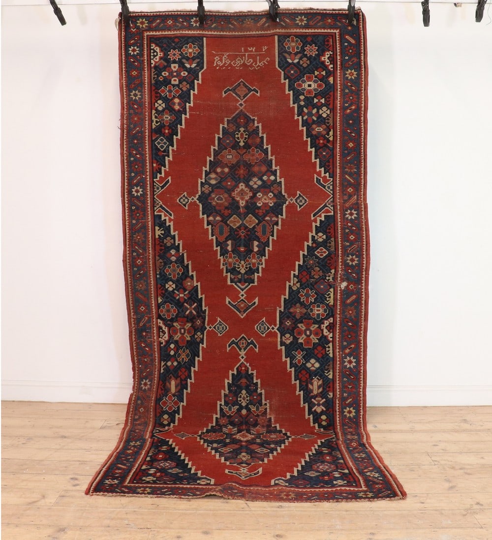 A Kazak carpet,: A Kazak carpet, A Kazak carpet, with two navy medallions, to a red field and navy floral borders, signed and dated, 312 x 130cm Condition Report: The pile is heavily worn. Repaired patches throughout.