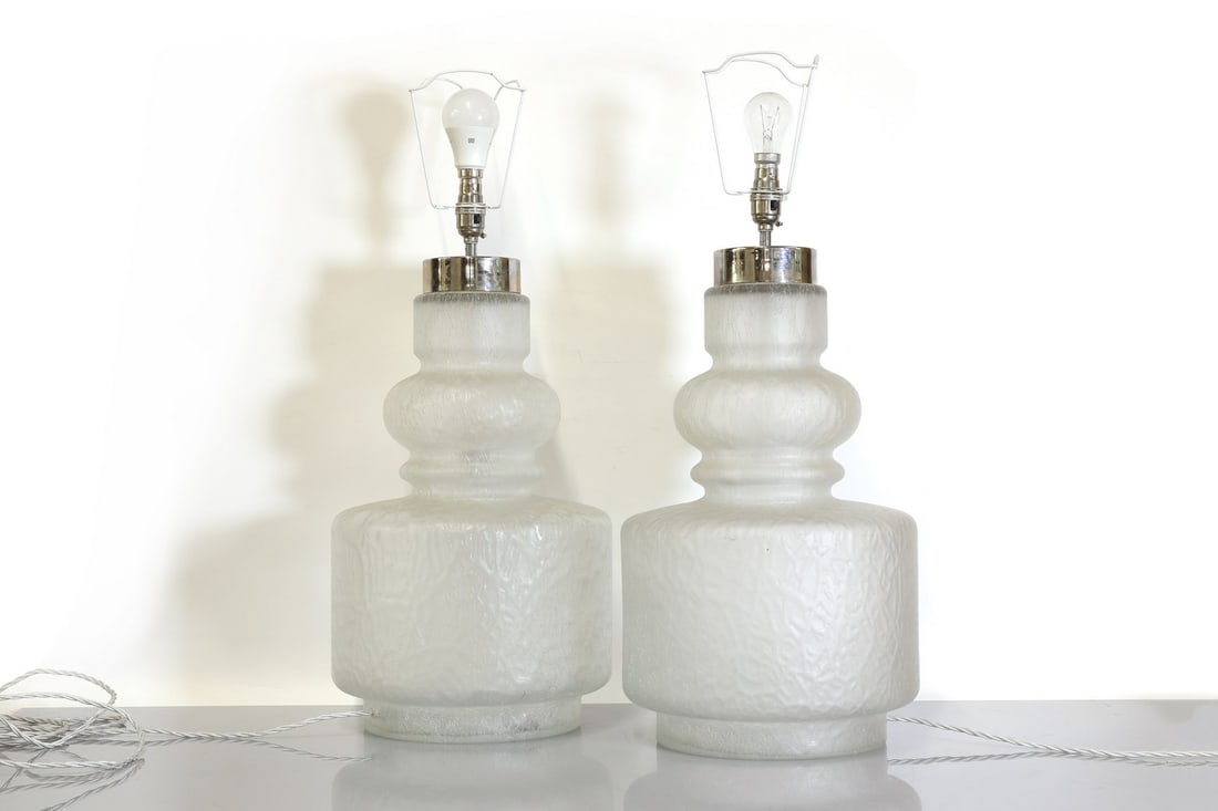 A pair of moulded glass table lamps,: A pair of moulded glass table lamps, A pair of moulded glass table lamps, 1970s, in the manner of Holmegaard, each frosted body of stepped form, with chromed fittings for two bulbs, with a pleated fab