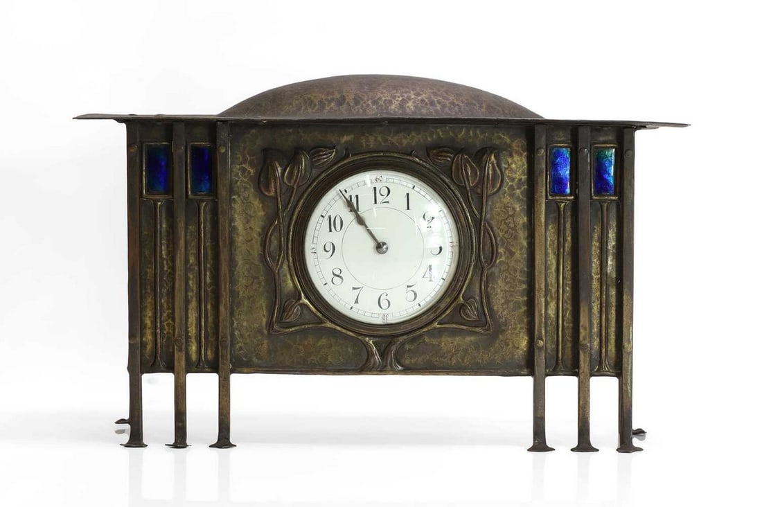A Glasgow School planished brass mantel clock,: A Glasgow School planished brass mantel clock, c.1900, attributed to George Walton (1867-1933), the round dial set in a repoussé surround of stylised foliage, raised to the front on six pilaster