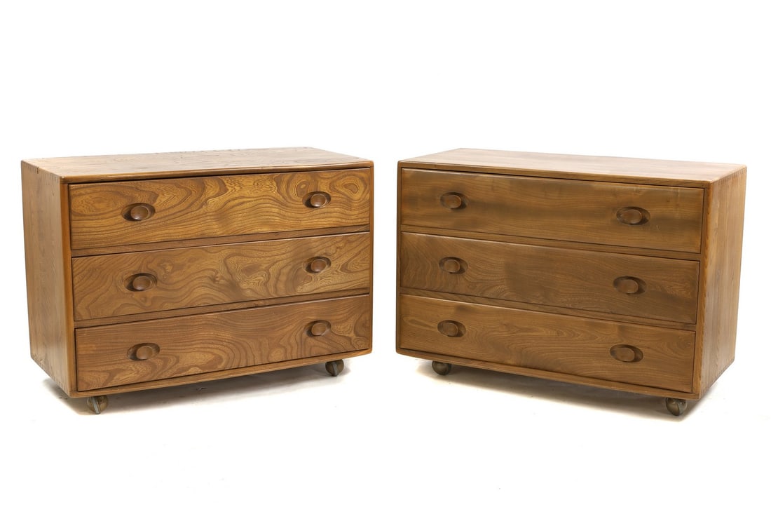 A pair of Ercol elm bedside chests,: A pair of Ercol elm bedside chests, each with three drawers, on castors, 91cm wide 44cm deep 66cm high together with a shaped wall mirror, 33cm wide 70cm high (3) Condition Report: With some wear. Som