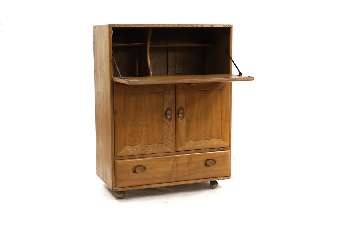An Ercol elm writing desk,: An Ercol elm writing desk, with a hinged writing surface, with shelf and division to the inside, over a cupboard and a drawer, on castors, 82.5cm wide 44cm deep 109.5cm high Condition Report: Used - g