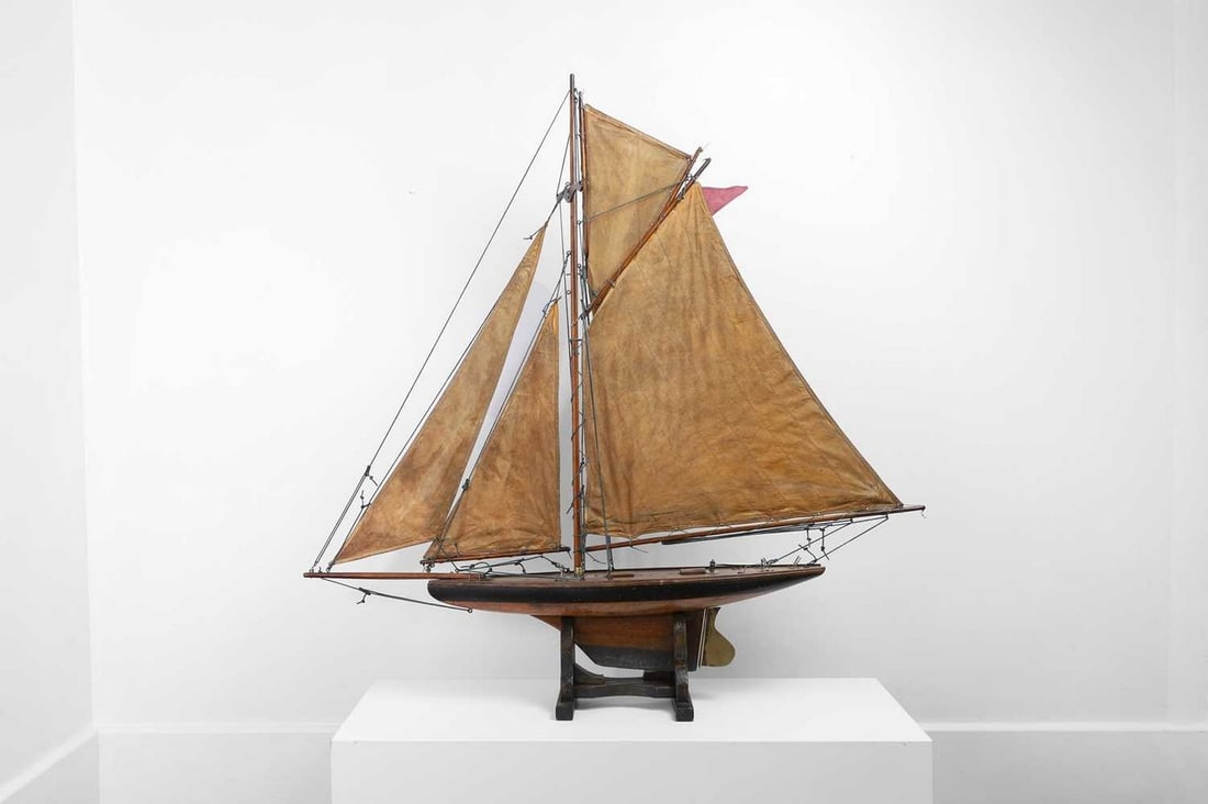 An Edwardian pond yacht,: An Edwardian pond yacht, early 20th century, with canvas sails, rope rigging and pinned hull with black-painted topside, on a wooden stand, 107cm wide 17cm deep 111cm high (2)