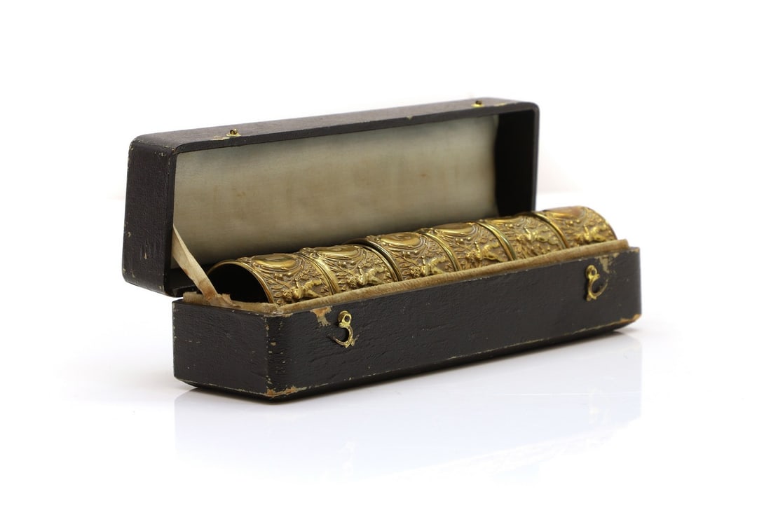 A set of six brass napkin rings,: A set of six brass napkin rings, early 20th century, each cast with a band of scrolling leaves and putti, both numbered, in a fitted case, 4.5cm diameter case 22cm wide Condition Report: Tarnished and