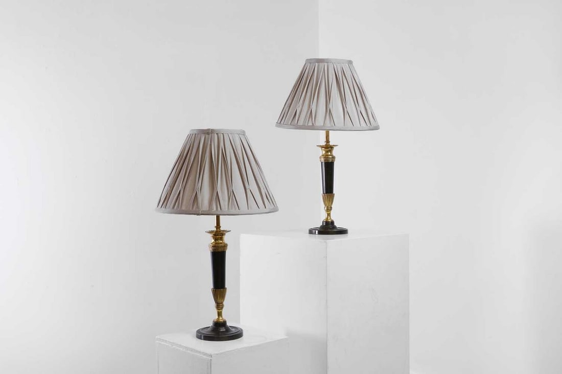 A pair of Empire-style gilt and patinated bronze lamps,: A pair of Empire-style gilt and patinated bronze lamps, 19th century and later, each in the form of a candlestick with a tapered column and raised on a circular foot, with a 12-inch grey pleated shade