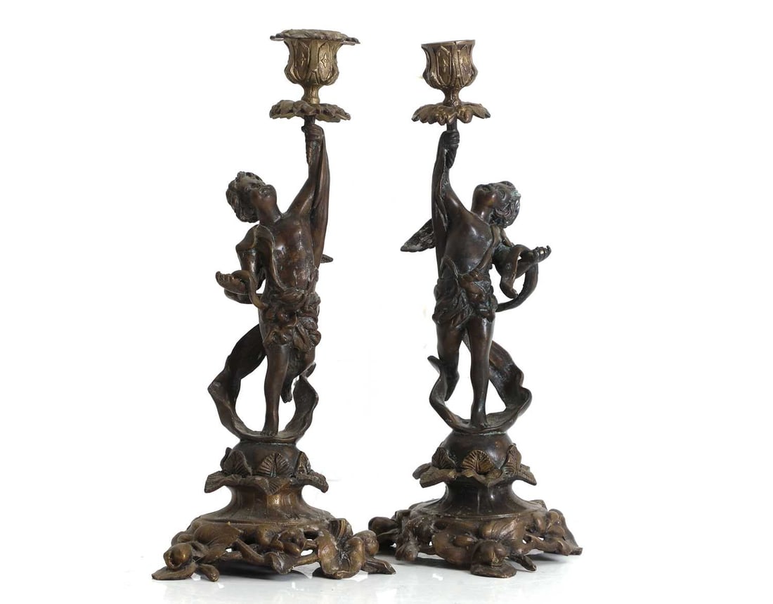 A pair of bronzed metal figural candlesticks, Late 19th to early 20th century: A pair of bronzed metal figural candlesticks, Late 19th to early 20th century each modelled as a winged putto standing on a foliate scroll base, supporting leaf cast drip pan 31.5cm high2