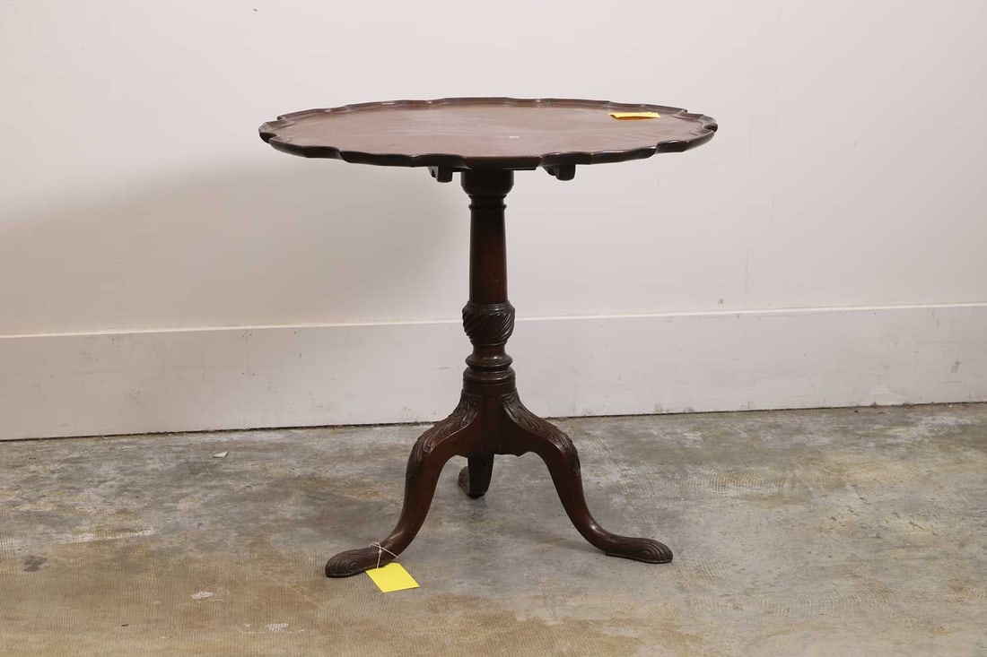 A mahogany tilt-top occasional table In the George III style: A mahogany tilt-top occasional table In the George III style The circular tilting top with a scalloped pie-crust edge above a baluster-turned column and a tripod base with carved cabriole legs and sc