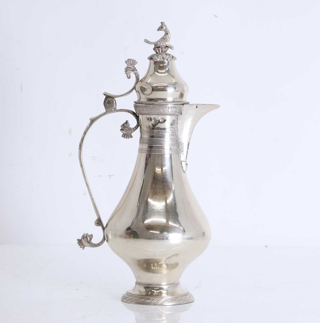 An Egyptian silver ewer,: An Egyptian silver ewer, marked Cairo, of baluster form on a raised foot, the s-scroll handle with bird head finials, the cover with applied bird finial, repeating floral design to the top, with
