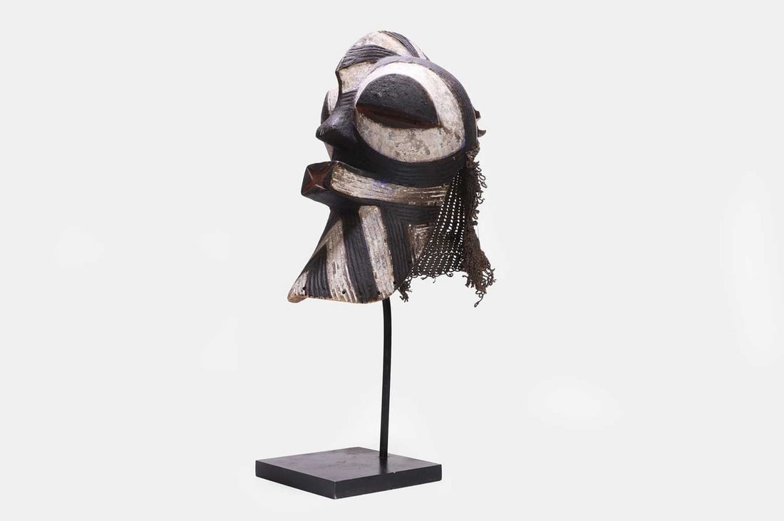 A Songye kifwebe mask, 20th century, Congolese,: A Songye kifwebe mask, 20th century, Congolese, carved wood with red, black and blue pigment, with a leather and fibre string cap, mounted on a black-painted metal stand (mask 21cm wide, 28cm deep, 36