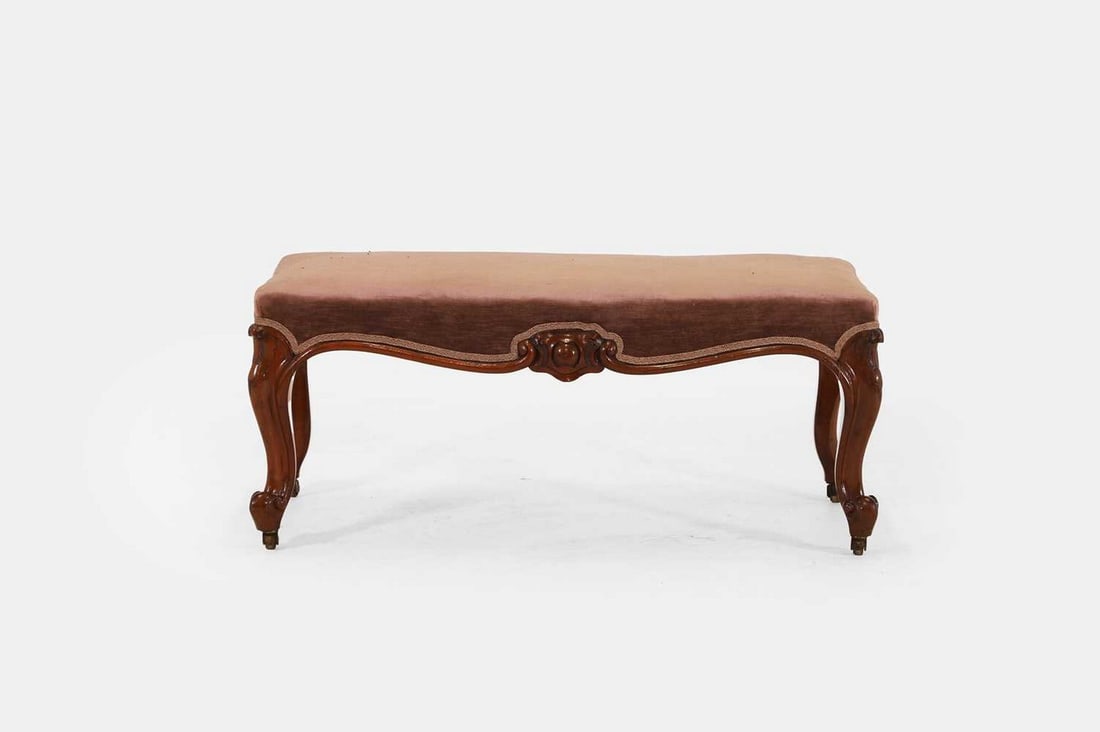 A Victorian mahogany stool, c.1870,: A Victorian mahogany stool, c.1870, with purple velvet upholstery and a shaped apron, raised on cabriole supports terminating in brass castors (103cm wide, 51cm deep, 45cm high) Condition Report: Ligh