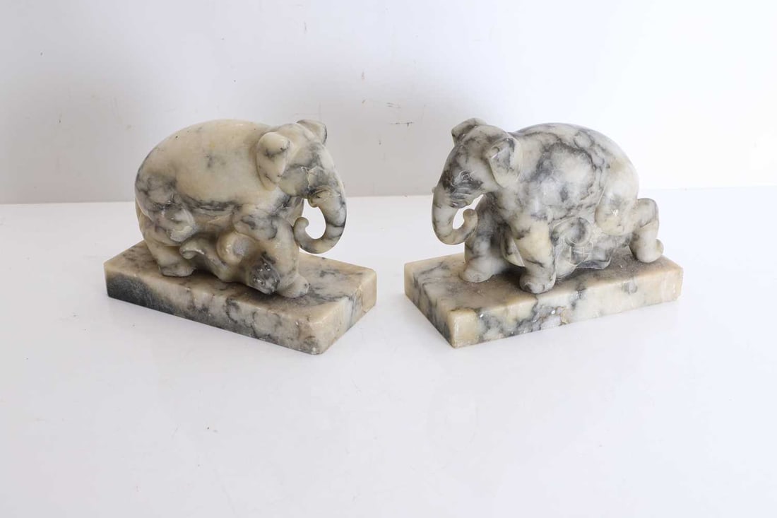 A pair of veined marble elephant bookends, 20th century,: A pair of veined marble elephant bookends, 20th century, each naturalistically carved as a kneeling elephant, modelled in variegated grey and cream marble, with dark veining, on integral plinth