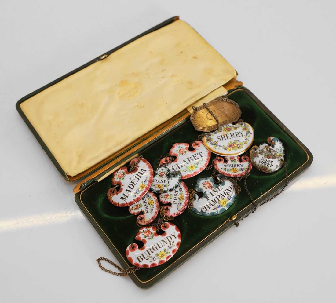 A collection of enamelled wine labels, 20th century,: A collection of enamelled wine labels, 20th century, each with chain suspensions, variously titled, and two other china examples, in a T Goode & Co, case, together with an Apsrey & Co