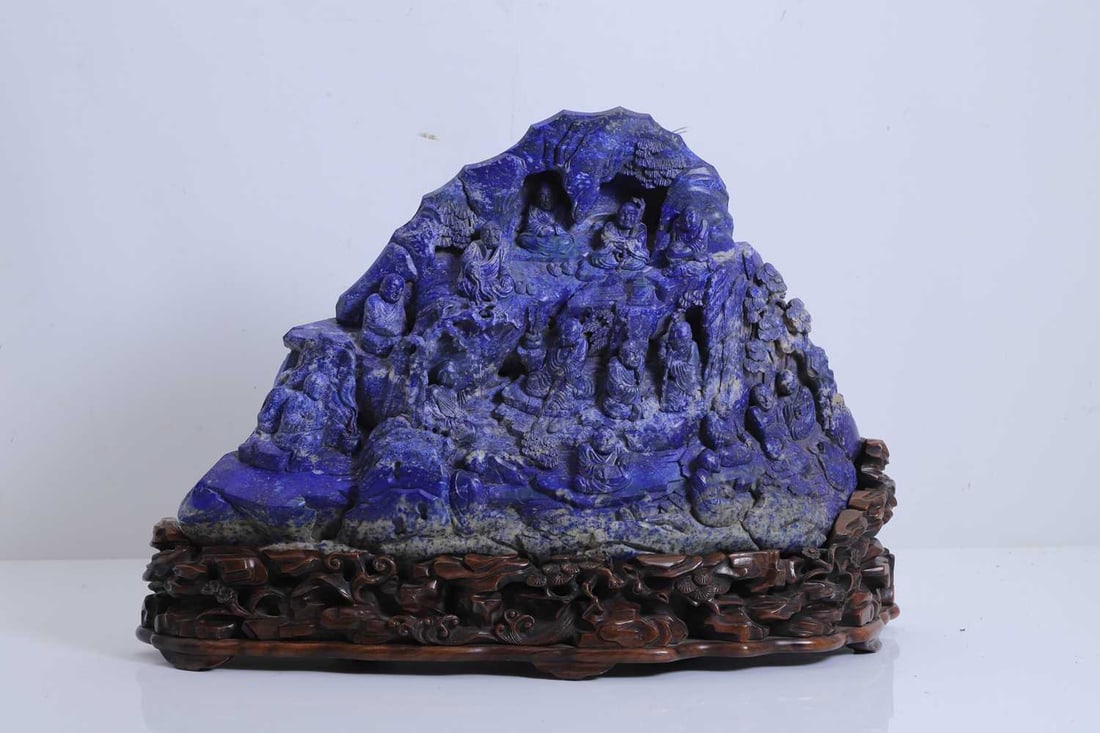 A Chinese large lapis lazuli figural group, 20th century,: A Chinese large lapis lazuli figural group, 20th century, carved as a mountain with sixteen monks amongst pine and foliage, mounted on a carved hardwood stand issuing pine, lingzhi and rockery 