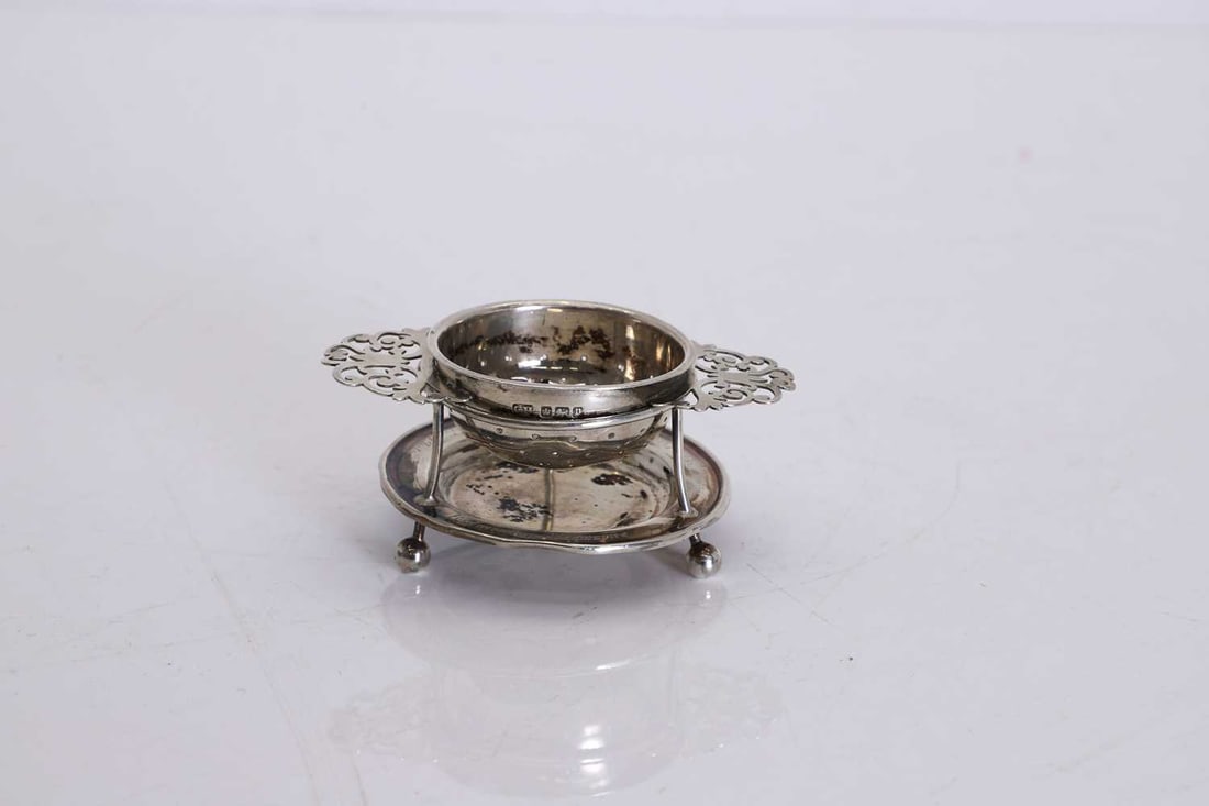 A silver tea strainer and stand,: A silver tea strainer and stand, by Harrison Brothers & Howson (George Howson), Sheffield 1907, the circular pierced strainer bowl with rolled rim, flanked by pierced foliate scroll handles,