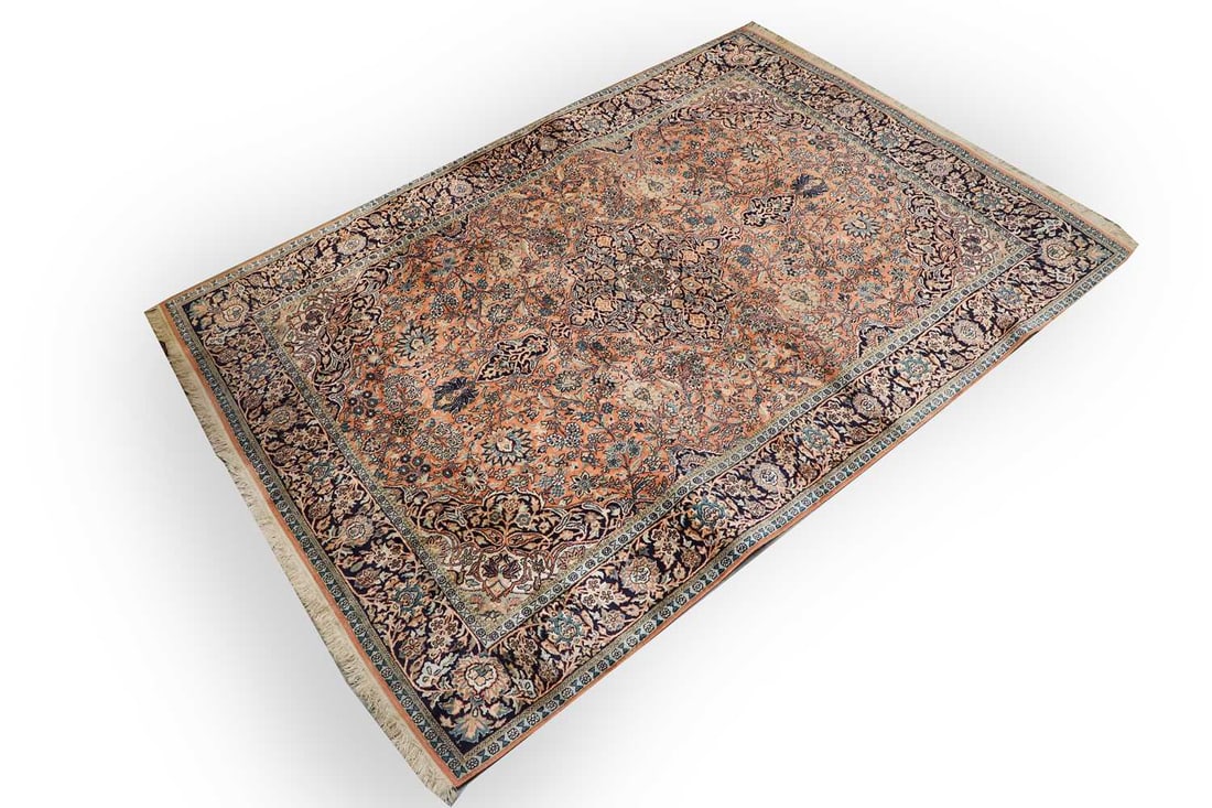 A silk Kirman carpet: A silk Kirman carpet of rectangular form, the coral field centred by a cusped medallion issuing an all-over design of scrolling vines, palmettes and flowering sprays, enclosed within a dark indigo bo