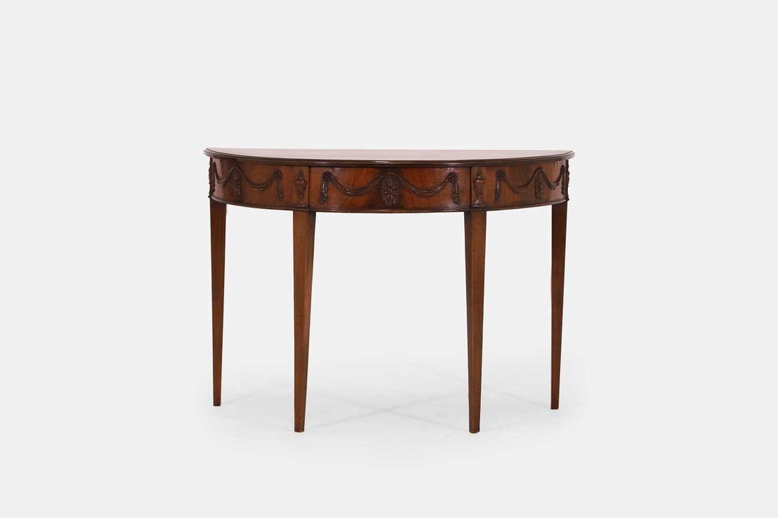 A George III-style mahogany side table, c.1890,: A George III-style mahogany side table, c.1890, with a plain demilune top above a frieze with carved swags, paterae and urns, raised on tapering square supports 100cm wide, 46.5cm deep, 70.5cm highCo