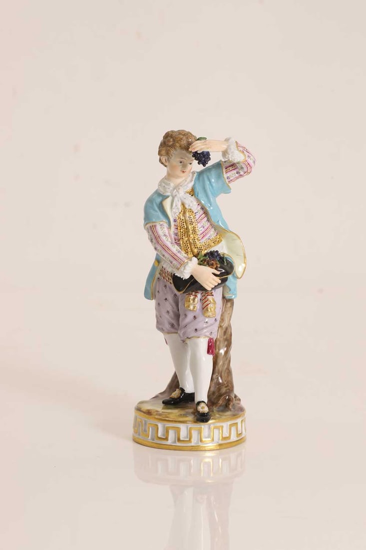 A Meissen porcelain figure of a grape seller, 19th century,: A Meissen porcelain figure of a grape seller, 19th century, imodelled as a young gentleman in 18th century dress, standing beside a tree stump, holding a bunch of grapes aloft in one hand and a