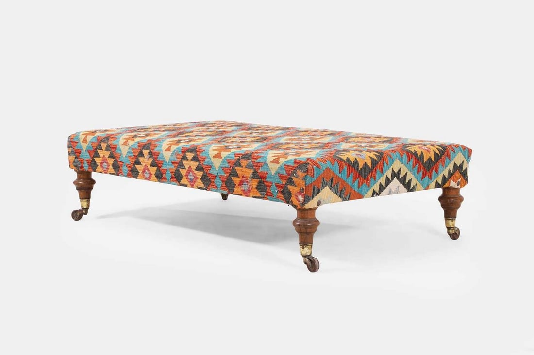 A large kilim ottoman stool, of recent manufacture with 19th-century legs and castors,: A large kilim ottoman stool, of recent manufacture with 19th-century legs and castors, the woollen upholstery decorated with repeating polychrome geometric motifs, raised on turned mahogany legs termi