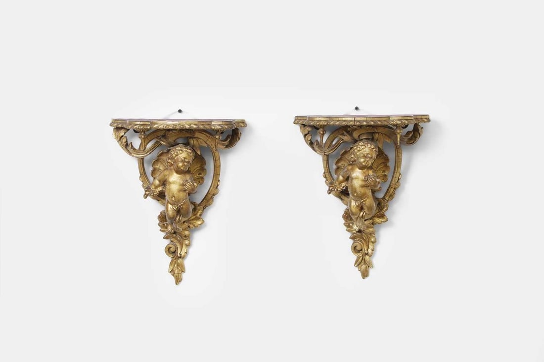 A pair of giltwood cherub wall brackets, 18th century, Continental,: A pair of giltwood cherub wall brackets, 18th century, Continental, each centred by a cherub surrounded by scrolling foliate, acanthus and rocaille detail, surmounted by a shaped purple velvet surface
