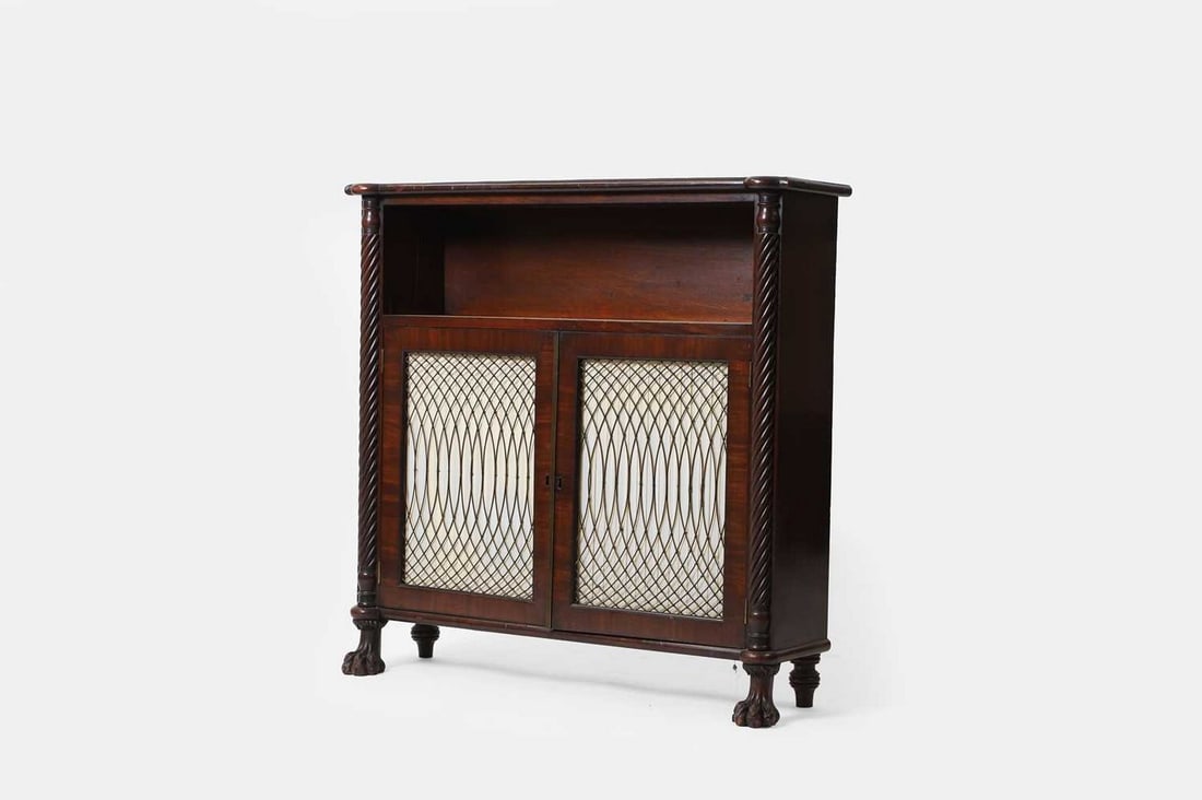 A small Regency mahogany bookcase, circa 1820, Irish, possibly Cork,: A small Regency mahogany bookcase, circa 1820, Irish, possibly Cork, the moulded top over an open bookshelf flanked by rope twist columns, above grille doors enclosing two shelves, raised on carved pa